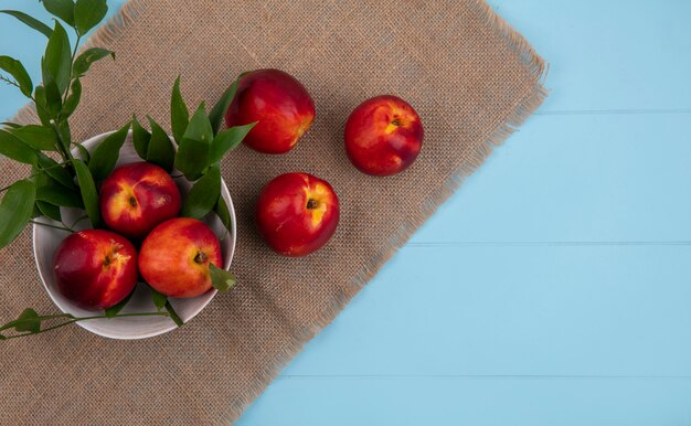 Top view of peaches in a bowl with leaf branches on a beige napkin on a light blue surface