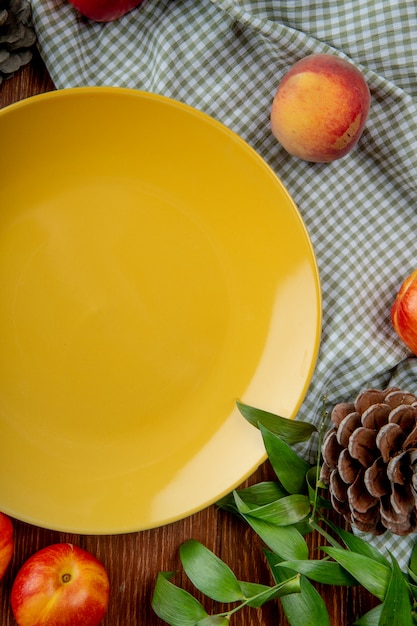 Вид сверху персики и шишка вокруг пустой тарелке на ткани на деревянной поверхности, украшенной листьями
