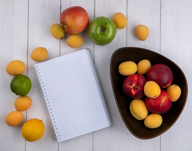 Top view of peaches with apricots in a bowl with apples and a notebook on a white surface