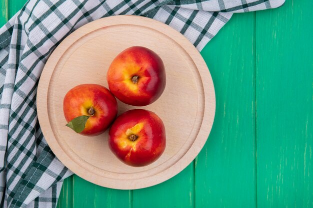 Top view of peaches on a wooden tray with a checkered towel on a green surface