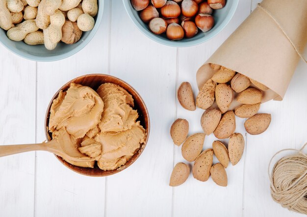 Top view of peanut butter in a bowl and almond in shell scattered on white background