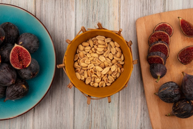 Free photo top view of peanuts on a bucket with black mission figs on a blue bowl with slices of black figs on a wooden kitchen board on a grey wooden wall