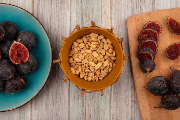 Top view of peanuts on a bucket with black mission figs on a blue bowl with slices of black figs on a wooden kitchen board on a grey wooden wall