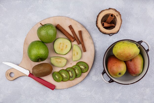 Top view pears in a saucepan with cinnamon  green apples and kiwi with a knife on a stand on a white background