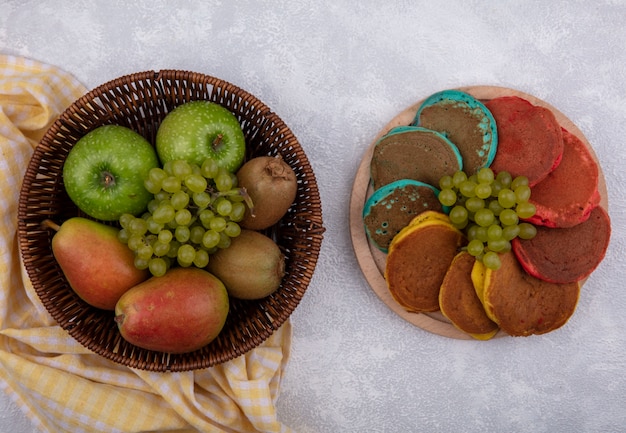 Top view pears with green apples  grapes and kiwi in a basket on a yellow checkered towel  with colored pancakes on a stand  on a white background