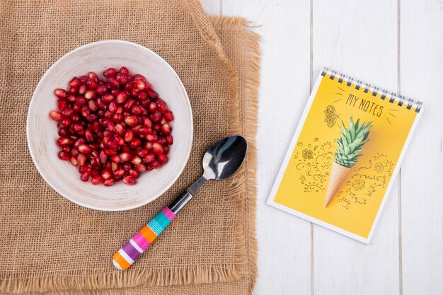 Top view of peeled pomegranate on a plate with a teaspoon on a beige napkin with a notebook on a white surface