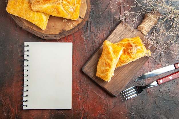 Top view of penovani khachapuri on wood board and on chopping board with knife and fork a notebook on dark red surface