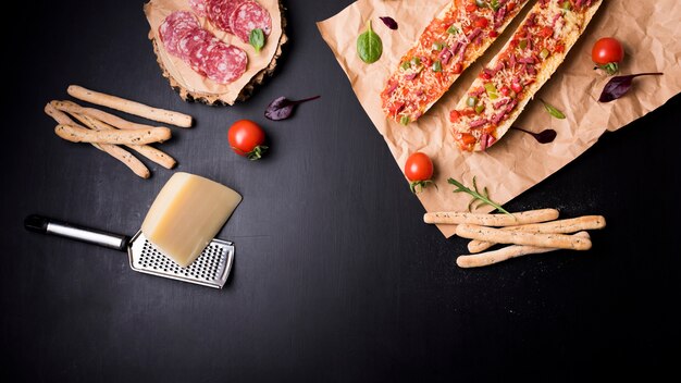 Вид сверху ломтиков пепперони; хлебные палочки; сыр; пицца с помидорами и багетом на коричневой бумаге над черной столешницей