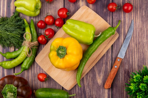 Top view of peppers on cutting board with dill tomatoes and knife on wooden surface