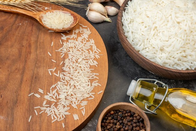 Top view of perfect long rice in a spoon in a brown bowl and on cutting board spikes garlics fallen oil bottle pepper on dark background