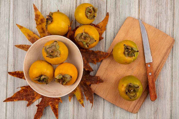top view of persimmon fruits on a bowl with leaves with persimmon fruits on a wooden kitchen board with knife on a grey wooden table