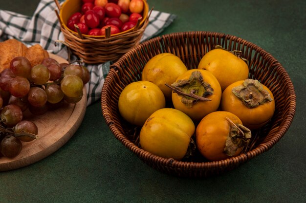 Top view of persimmon fruits on a bucket with grapes on a wooden kitchen board on a checked cloth on a green background