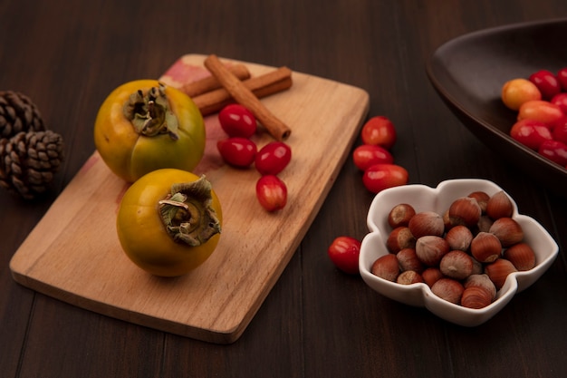 Top view of persimmon fruits on a wooden kitchen board with cinnamon sticks with hazelnuts on a bowl with cornelian cherries isolated on a wooden surface