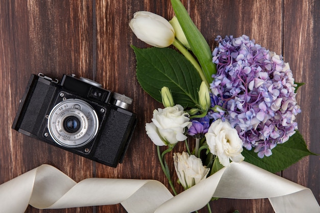 Top view of photo camera and flowers with white ribbon on wooden background