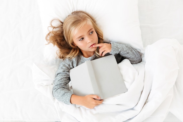Top view photo of thoughtful little girl lying in bed with gray book, looking aside
