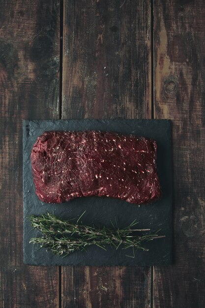 Top view piece of raw meat on black stone pad with rosemary, all on aged wooden table