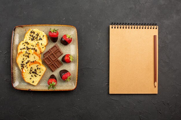 어두운 테이블에 갈색 연필이 있는 노트북 옆에 초콜릿과 딸기가 있는 식욕을 돋우는 케이크 조각