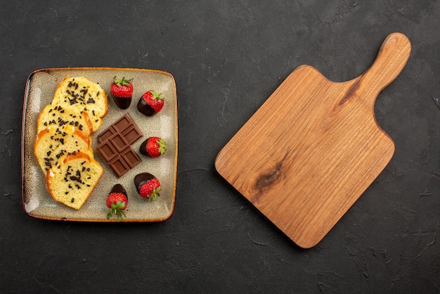 Top view pieces of cake pieces of cake with chocolate and strawberries and brown kitchen board on dark table