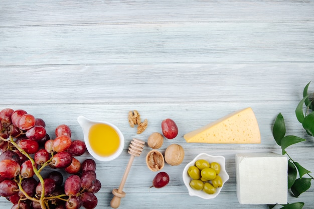 Top view of pieces of cheese with honey, fresh grape, pickled olives and walnuts on grey wooden table with copy space