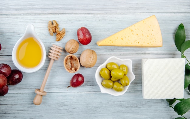 Top view of pieces of cheese with honey, fresh grape, pickled olives and walnuts on grey wooden table