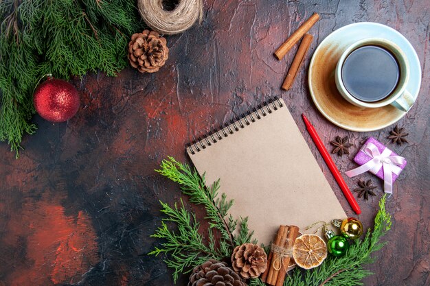 Top view pine tree branches and pinecones a notebook red pen dried lemon slices straw thread cup of tea anises on dark red surface with free place