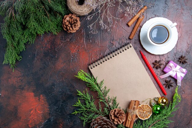 Top view pine tree branches and pinecones a notebook red pen dried lemon slices straw thread cup of tea star anises on dark red surface with free place