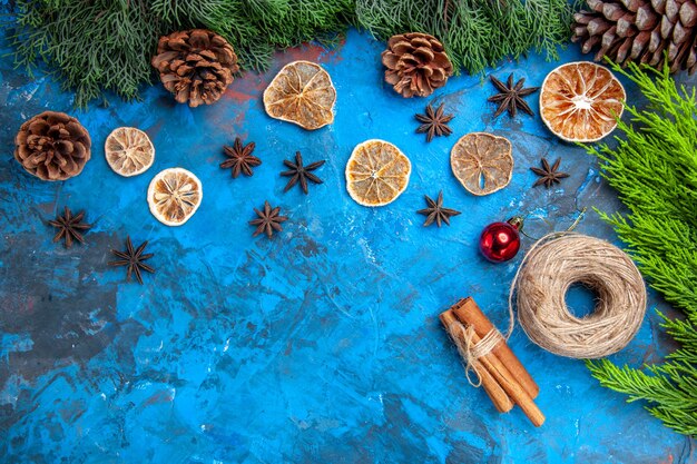 Top view pine tree branches pinecones straw thread cinnamon sticks dried lemon slices anise seeds on blue-red surface