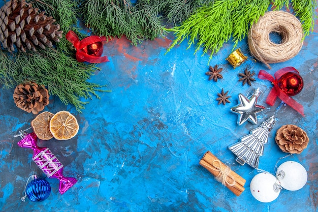 Вид сверху сосновые ветки сосновые шишки соломенная нить рождественские елочные игрушки семена аниса палочки корицы сушеные дольки лимона на сине-красной поверхности