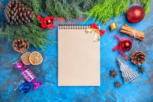Top view pine tree branches pinecones xmas tree toys anise seeds dried lemon slices a notebook on blue-red background
