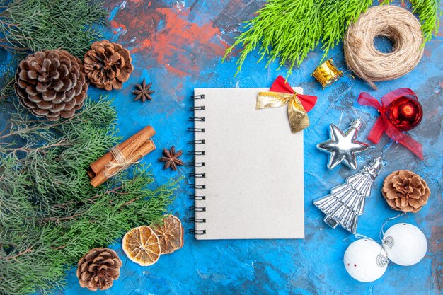 Top view pine tree branches with cones cinnamon sticks anise seeds dried lemon slices straw thread xmas tree toys a notebook on blue-red background