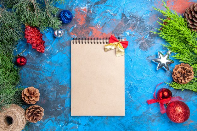 Top view pine tree branches with pinecones and colorful xmas tree toys straw thread a notebook on blue-red background