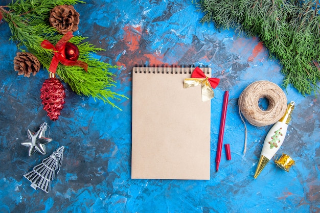 Top view pine tree branches with pinecones hanging ornaments notebook red pen straw thread on blue-red surface