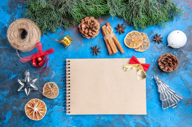 Top view pine tree branches xmas tree toys straw thread cinnamon sticks dried lemon slices anise seeds a notebook on blue-red background