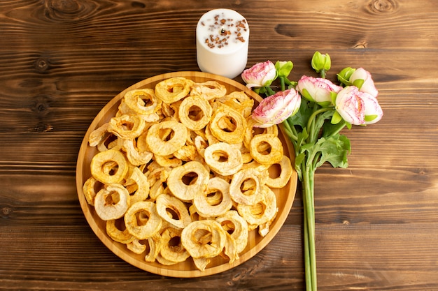 A top view pineapple dried rings inside plate dried fruits sour tasty unique taste along with pink flowers on the brown wooden desk fruits exotic dry