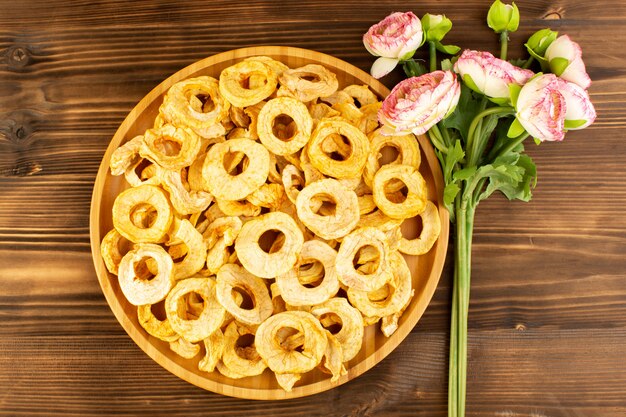 Вид сверху ананасовых сушеных колец внутри тарелки сухофруктов кислого вкусного неповторимого вкуса вместе с розовыми цветами на коричневом деревянном столе фруктов экзотических сухих