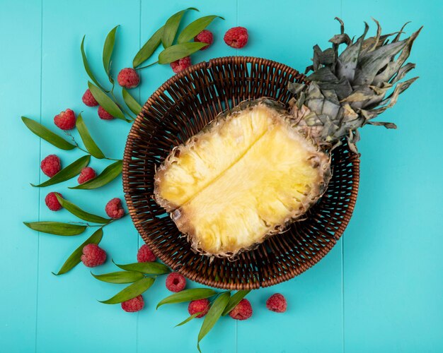 Top view of pineapple half in basket with pattern of raspberries and leaves on blue surface