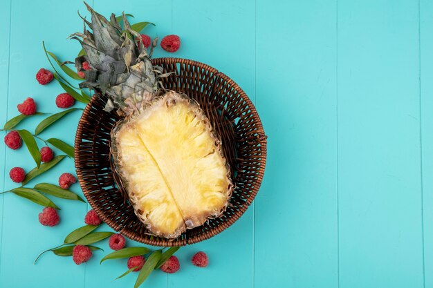 Top view of pineapple half in basket with pattern of raspberries and leaves on blue surface