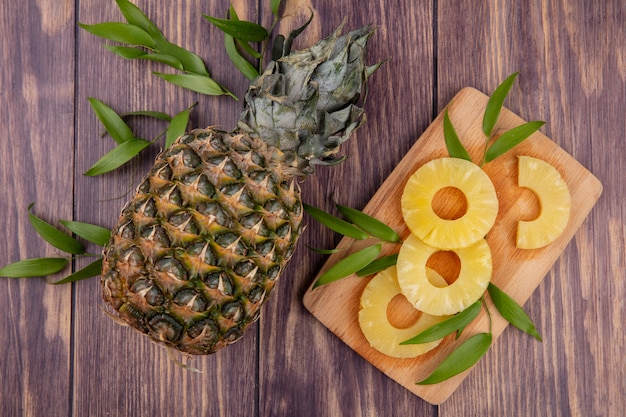 Top view of pineapple and pineapple slices with leaves on cutting board and wooden surface