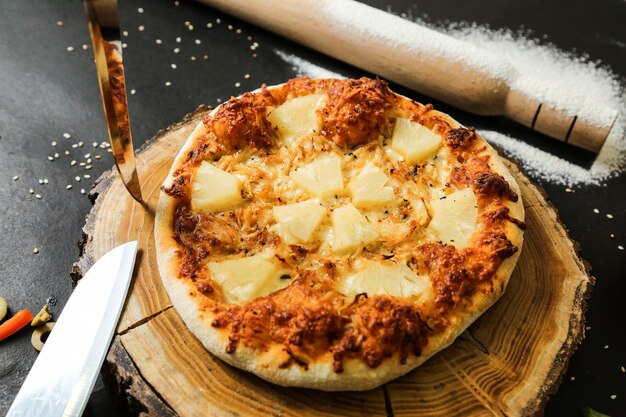 Top view pineapple pizza on stand with knives rolling pin and flour on black table