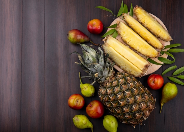 Top view of pineapple slices in plate with whole one and peach plum on wooden surface