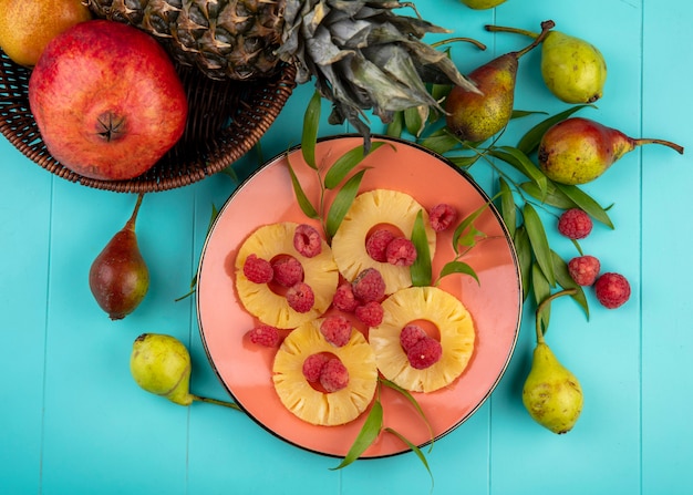Free photo top view of pineapple slices and raspberries in plate with basket of pineapple pomegranate and peaches with leaves around on blue surface