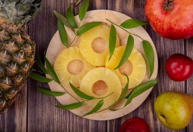 Top view of pineapple slices with leaves on cutting board and pineapple pomegranate peach plum on wooden surface