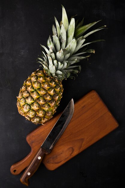 Top view of pineapple with knife on cutting board on black surface
