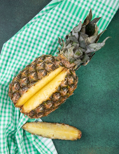 Top view of pineapple with one piece cut out from whole fruit on plaid cloth and green surface