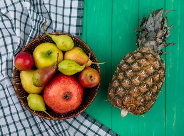 Top view of pineapple with pomegranate apple peach plum in basket on plaid cloth and green surface