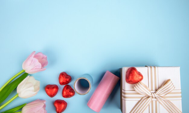 Top view of pink color tulips heart shaped chocolate candies wrapped in red foil, gift box and roll of colorful paper on blue table with copy space