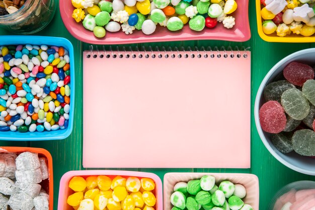 Free photo top view of a pink sketchbook and bowls with various colorful candies on green background