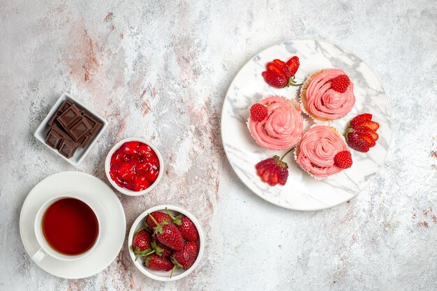 Top view of pink strawberry cakes with cream and cup of tea on white surface