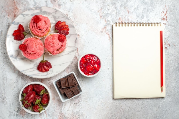 Top view of pink strawberry cakes with jam and chocolate bars on white surface