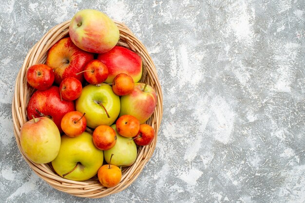 회색 테이블의 왼쪽에 빨간색과 노란색 사과와 자두가있는 상위 뷰 플라스틱 바구니